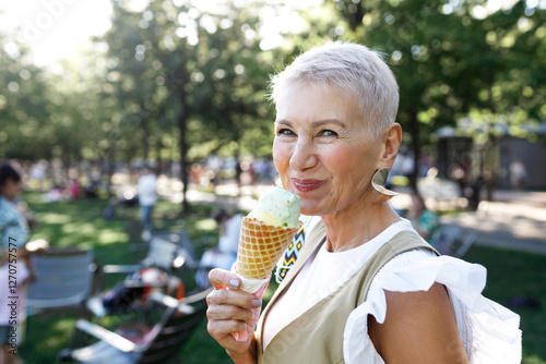 Side view outdoor portrait of smiling senior female with short stylish haircut eating tasty delicious pistachio ice cream, enjoying as kid, during her evening promenade in public park
