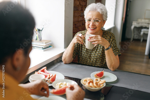 Two senior female best friends drinking tea and having nice friendly conversation with each other sitting at table with sweets, caucasian woman laughing looking at her african american bestie photo