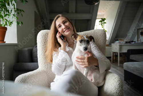 Smiling woman talking on smart phone and sitting with dog at home photo