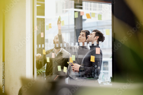 Businesswoman sticking adhesive note on glass wall and discussing strategies with coworkers in office photo