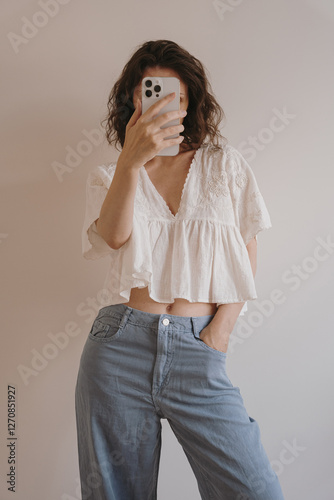 Fotografie Woman in White Top and Denim Taking Mirror Selfie with Smartphone in Simple Room