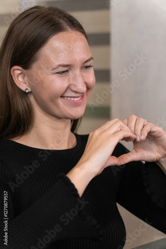 Smiling young woman making heart shape with hands while using laptop in modern kitchen, expressing love and affection in online communication. Vertical photo