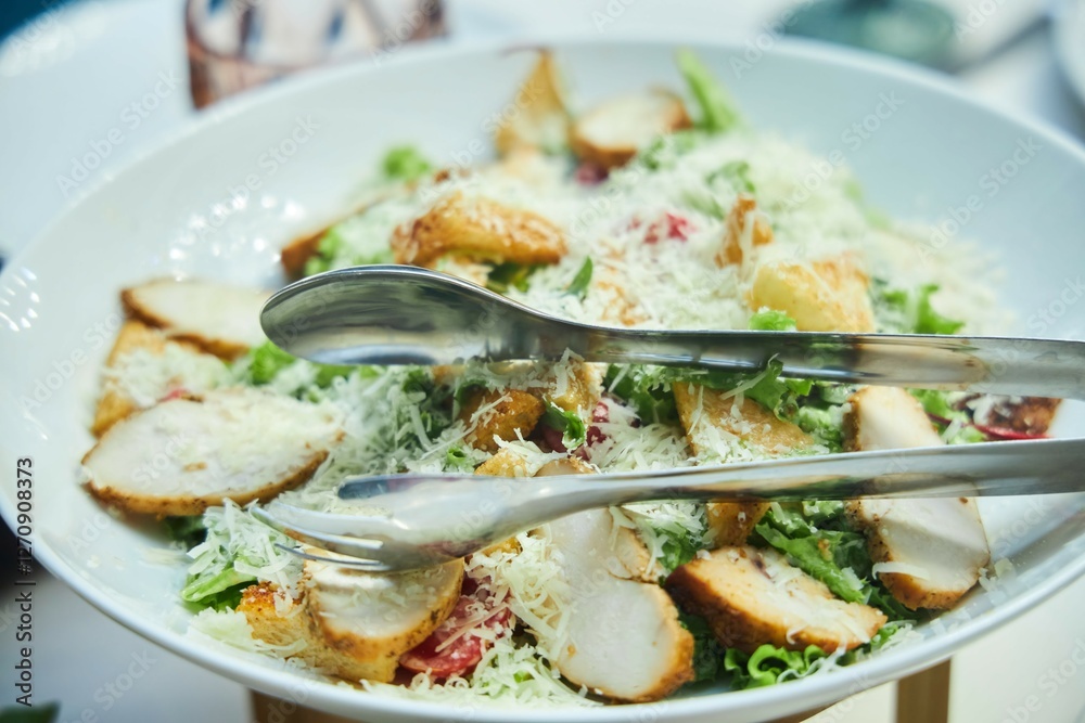 On a white plate there is a salad with chicken, a fork and a knife. The salad is decorated with cheese and lettuce leaves. Caesar salad with chicken