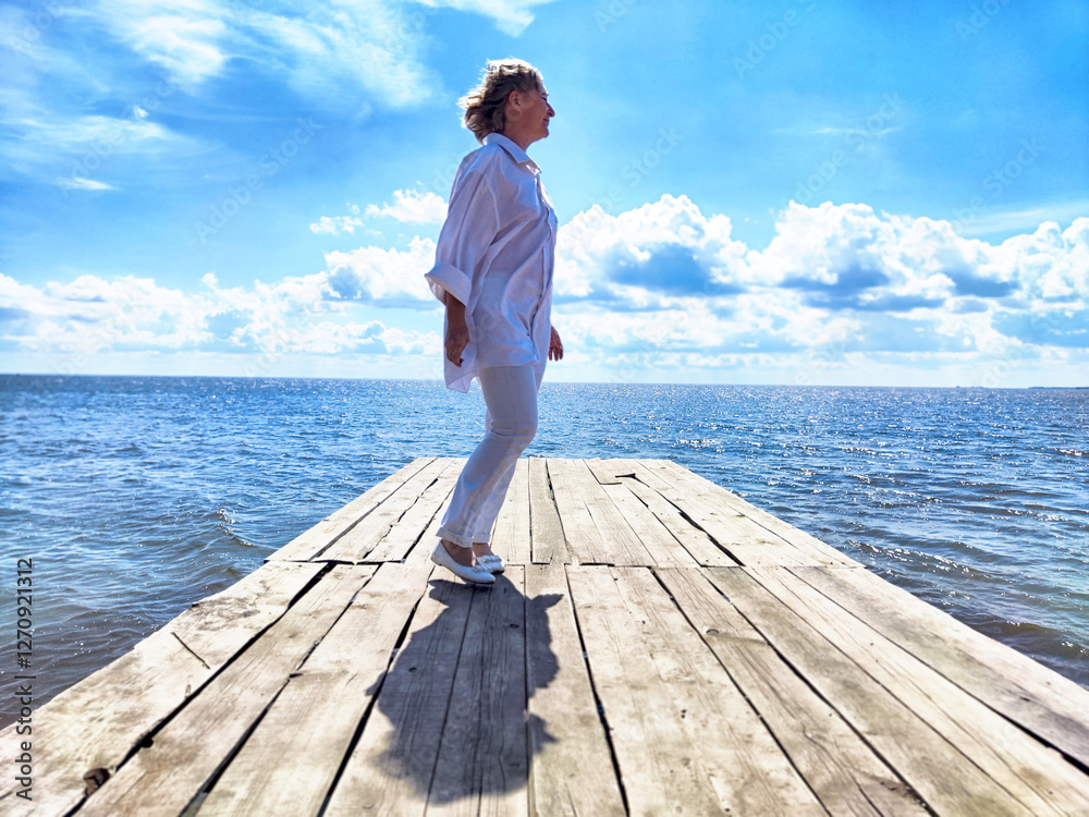 Blonde middle-aged woman enjoying a serene moment on a wooden pier by the sea under a bright blue sky