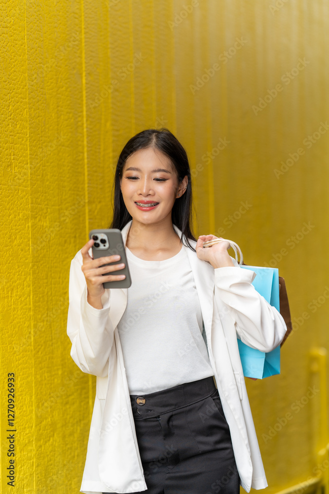 Smiling Shopper: A young woman, smiling and holding shopping bags, leans against a yellow wall while using her smartphone, encapsulating the modern shopping experience.