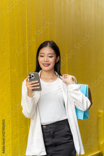 Smiling Shopper: A young woman, smiling and holding shopping bags, leans against a yellow wall while using her smartphone, encapsulating the modern shopping experience.