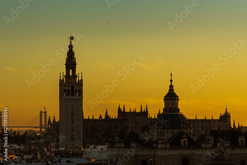 The iconic Giralda tower and Seville Cathedral form a dark silhouette against a glowing orange and green sunset sky in Spain
