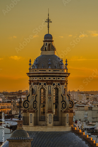 A decorative tiled church dome with a cross rises above the Seville rooftops against a vivid orange sunset sky in Spain