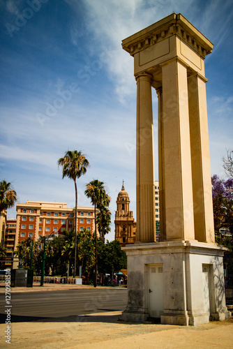 A tall stone monument column stands in a sunny urban plaza with Malaga Cathedral dome and palm trees visible behind in Spain