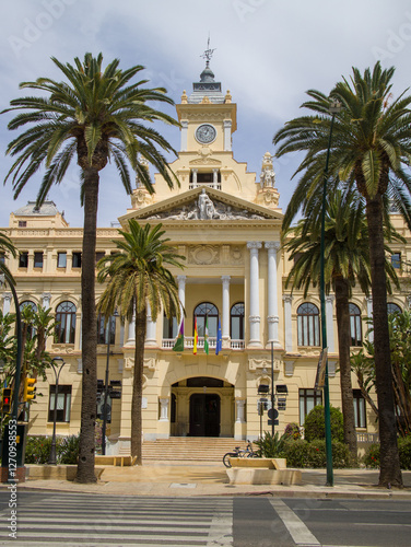 The ornate yellow facade of the Malaga City Hall stands between tall palm trees with Spanish flags flying in Malaga Spain