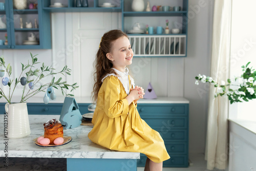 Happy child girl praying, hands folded in prayer concept for faith, spirituality and religion. Smiling little blond girl in a yellow dress on kitchen with easter cake, flowers, easter eggs, bunny. 