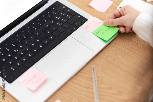 A person places colorful sticky notes with meeting schedules on a laptop keyboard while organizing tasks in a bright workspace with a wooden desk.