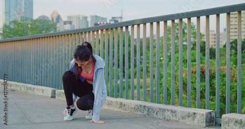 A female runner in sportswear bends over, holding her leg in pain after an accident or muscle strain during an outdoor workout on a bridge.
