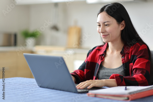 Satisfied asian student writing on laptop in the kitchen photo