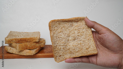 A hand holding a slice of whole wheat bread with a pile of bread beside it and a white background (isolated white). photo