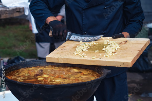 The chef adds garlic chopped on a cutting board to the meat soup with a knife. Boiling fresh food from vegetables and beef in a large cauldron. Catering and small business