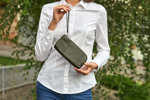 Obraz na płótnie Woman in white shirt holding olive green leather purse near outdoor foliage in afternoon sun, fashion accessory concept w salonie