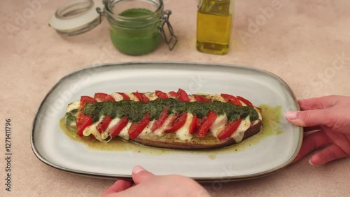 Baked eggplant with mozzarella cheese, tomatoes and pesto sauce. The woman puts a plate on the table. Traditional homemade seasonal dish. Grilled stuffed eggplant. Hearty summer dish. Selective focus.