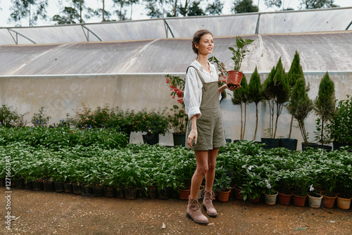 Young woman in a gardening outfit holding a potted plant, surrounded by greenery in a greenhouse, exuding joy and passion for gardening
