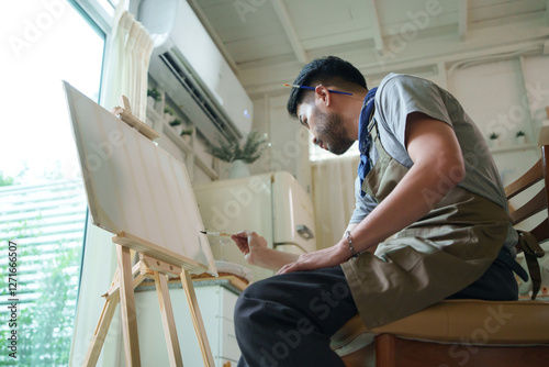 Asian man focused on painting at easel in bright, serene room, surrounded by art supplies