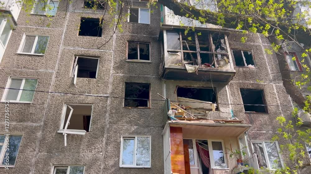 A damaged residential building with broken windows, a destroyed balcony, and visible debris. The facade shows signs of fire and destruction. Green leaves on a tree contrast with the ruined structure.
