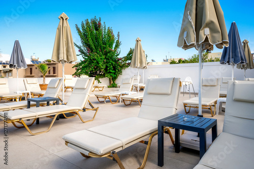 Luxury rooftop sunbathing area with white loungers, beige umbrellas, and a modern outdoor setting. Clear blue sky, greenery, and urban buildings create a relaxing vibe