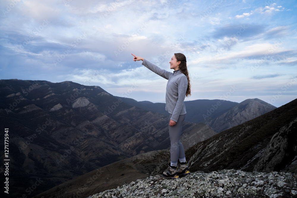Beautiful woman traveler standing and pointing towards the sky in the mountains at the twilight sunset. Female hiker direction her finger the way wearing sportswear. Adventure, exploration, hiking.