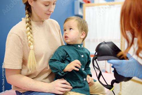 Toddler boy with mom are at appointment with a pediatric neurologist. Neurology doctor is establishes contact and trust with the child. Doctor prepares to make positional testing of patient using VNG photo