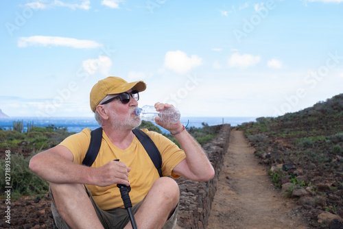 Senior man in hat and yellow t-shirt drinks from bottle water sitting outdoors on seaside excursion. Relaxed caucasian man enjoying his retirement by leading a healthy lifestyle photo