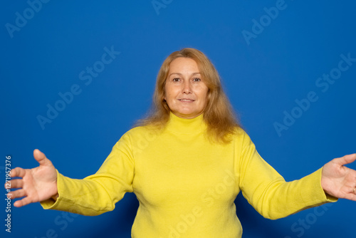 Caucasian woman in her 40s wearing a yellow sweater with her arms open in a welcoming gesture, isolated on blue studio background.