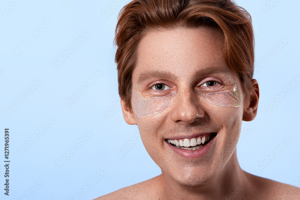 Fototapeta premium young man with sensitive skin with freckles and red hair wears hydrogel patches under his eyes, male skin care, feminine man concept isolated on blue background