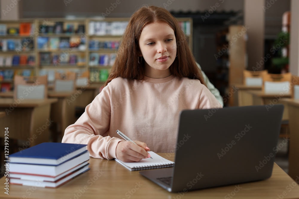 Girl taking notes while using laptop at wooden desk in public library Girl taking notes while using laptop at wooden desk in public library