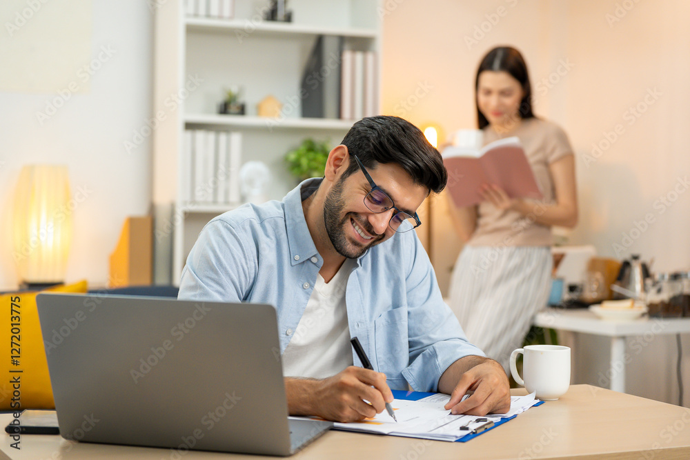 Young businessman working at home office on laptop computer, finishing his online work, while his girlfriend and roommate sitting behind on sofa relaxing and waiting. Freelance business. couple. Young businessman working at home office on laptop computer, finishing his online work, while his girlfriend and roommate sitting behind on sofa relaxing and waiting. Freelance business. couple.