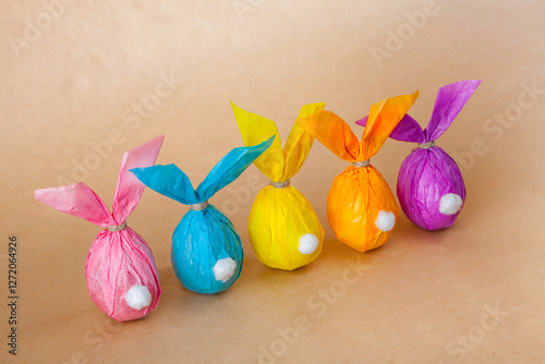 Five eggs wrapped in colored paper resemble adorable bunnies with ears and white tails. cute bunnies stand on a light brown surface, creating a charming Easter scene.