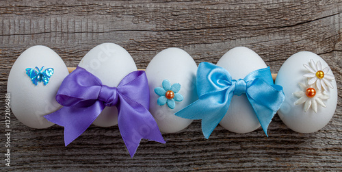 Several eggs are on the wooden table prepared for the Easter holiday. Each egg is decorated with colorful beads, as well as butterflies and stars made of fabric. Some eggs are adorned with blue ribbon