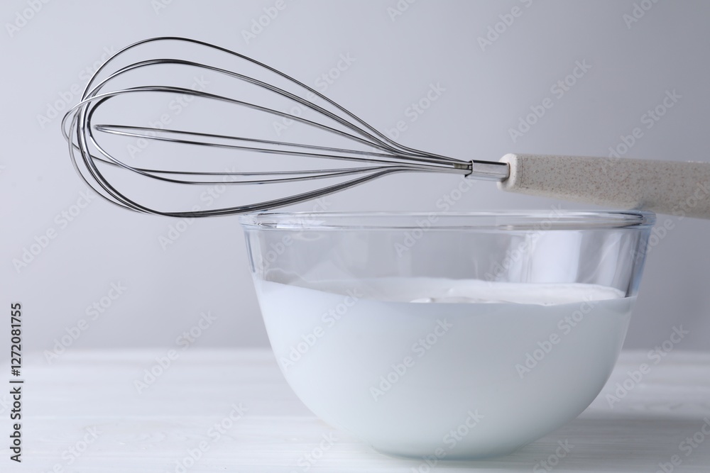 Whisk and bowl of whipped cream on white wooden table, closeup