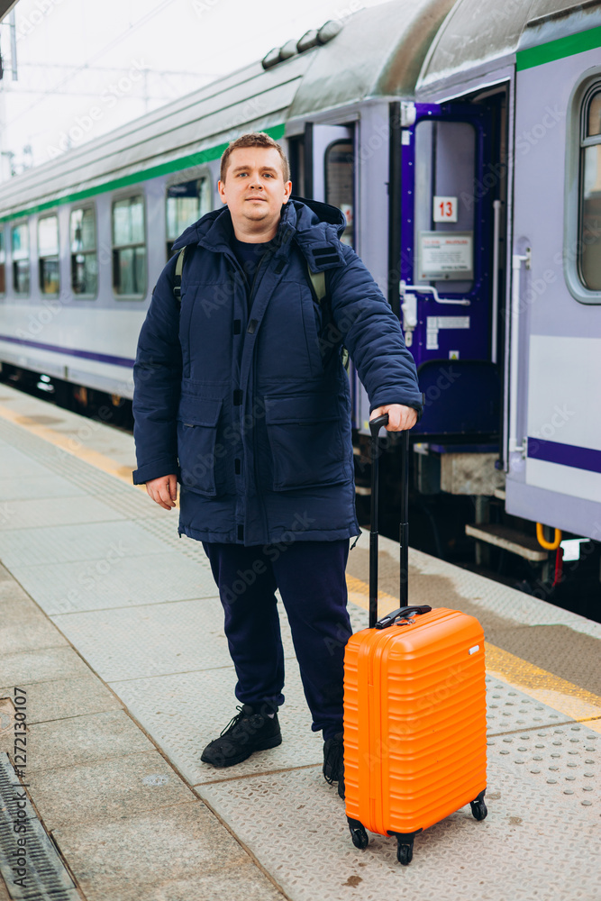 A young man with a small orange suitcase waits for a train at the train station for traveling. Lifestyle, people, adventure and tourism concept. High quality photo.
