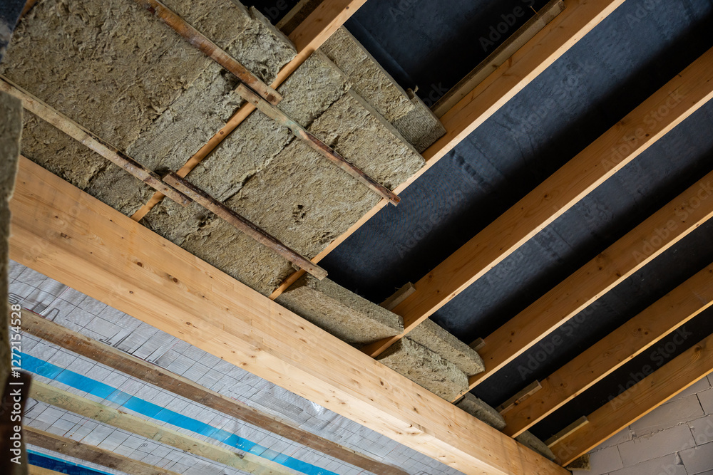 Exposed wooden beams and insulation material are visible in a building under construction, with a black vapor barrier on the ceiling section. Exposed wooden beams and insulation material are visible in a building under construction, with a black vapor barrier on the ceiling section.