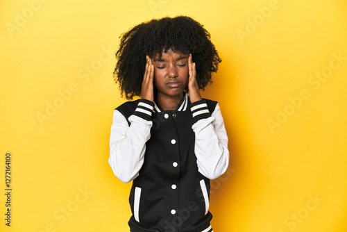 Teen girl in baseball jacket, yellow studio background touching temples and having headache. photo