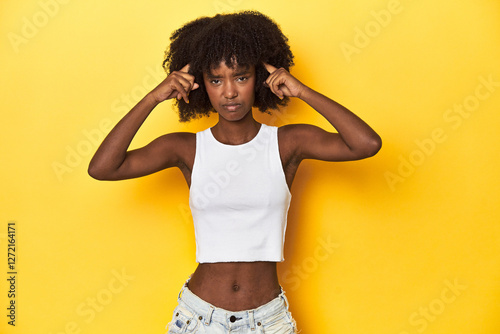Teen girl in white tank top, yellow studio background focused on a task, keeping forefingers pointing head.