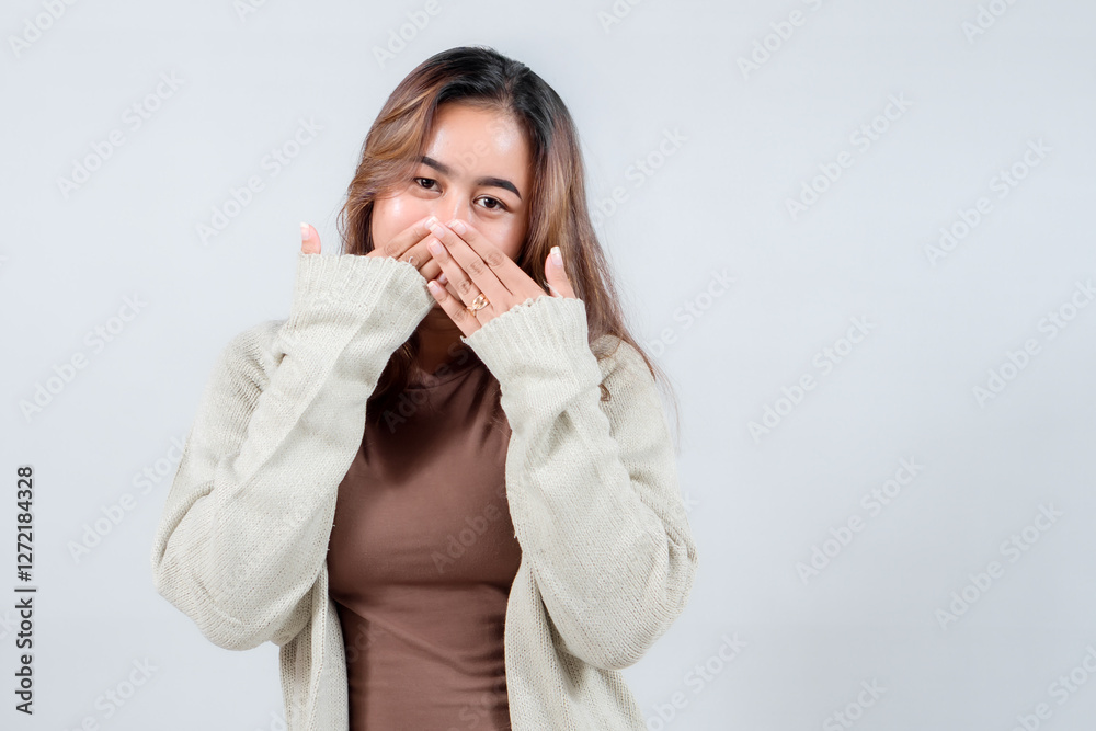 A young woman in a cozy sweater covers her mouth with her hands, expressing secrecy, surprise, or shyness against a neutral background A young woman in a cozy sweater covers her mouth with her hands, expressing secrecy, surprise, or shyness against a neutral background