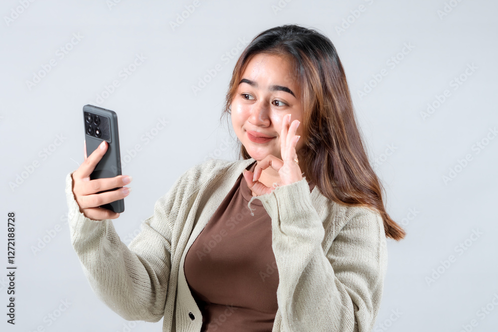 A young woman smiles and waves at her smartphone, expressing friendliness and warmth while engaging in a video call against a neutral background A young woman smiles and waves at her smartphone, expressing friendliness and warmth while engaging in a video call against a neutral background