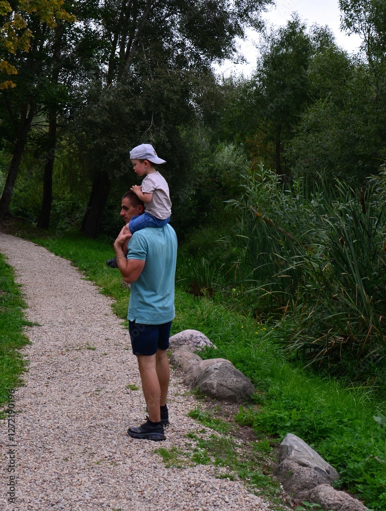Father carrying his boy on neck outside in summer. Son sits on his dads neck and looks around. Fun family time outside. Father carrying his boy on neck outside in summer. Son sits on his dads neck and looks around. Fun family time outside.