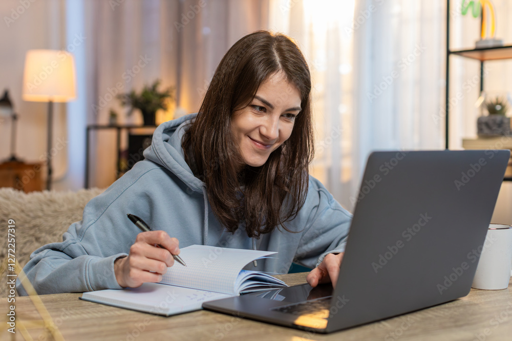 Young Caucasian woman watching online course on laptop sitting at table on sofa couch taking notes in notebook diary. Thoughtful female freelancer in casual clothes working from home in living room. Young Caucasian woman watching online course on laptop sitting at table on sofa couch taking notes in notebook diary. Thoughtful female freelancer in casual clothes working from home in living room.