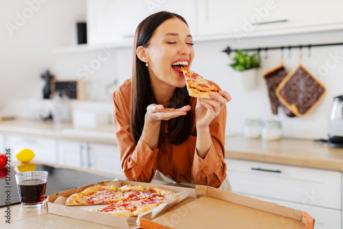 The joy of pizza. Excited woman enjoying tasty pizza, holding and biting slice with closed eyes, sitting at table in kitchen. Cheat meal, delivery service concept