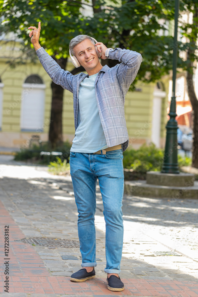 Happy relaxed overjoyed gray-haired man in wireless headphones listening disco music dancing outdoors. Cheerful guy in casual clothes walking in urban city street park. Town lifestyles. Vertical. Happy relaxed overjoyed gray-haired man in wireless headphones listening disco music dancing outdoors. Cheerful guy in casual clothes walking in urban city street park. Town lifestyles. Vertical.