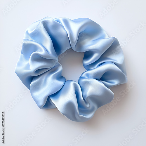 A light blue scrunchie with a soft, silky texture rests on a clean, neutral white background, casting a gentle shadow.