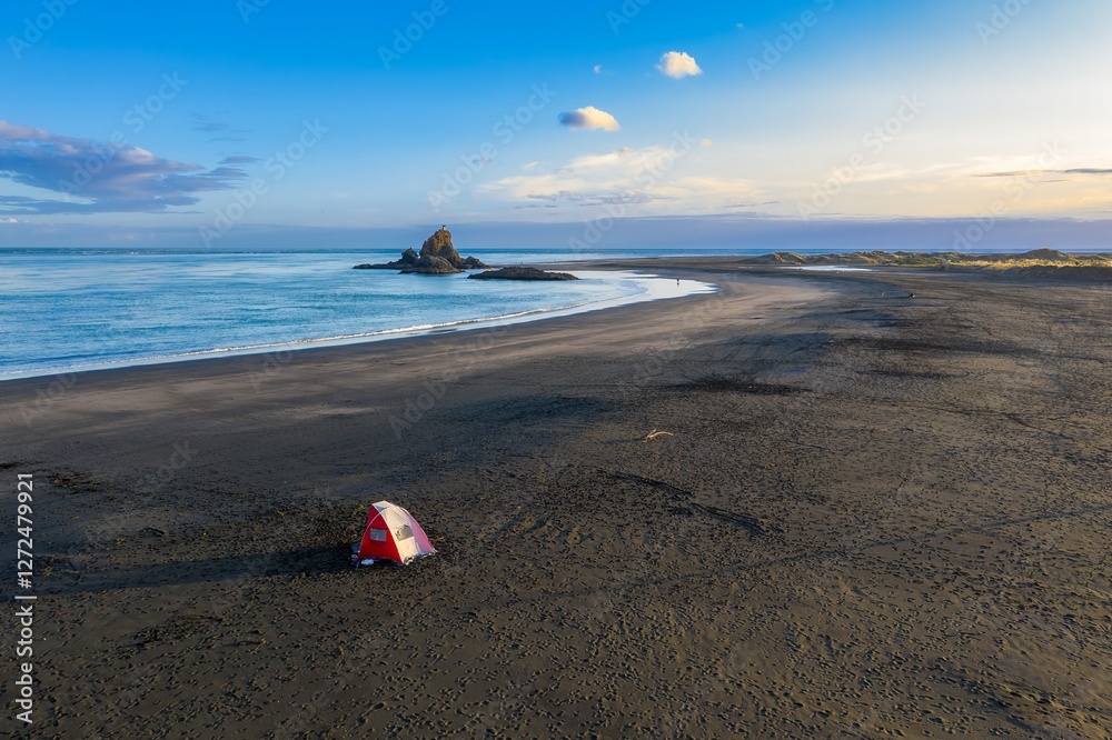Obraz premium Empty beach campsite at sunset on Whatipu. Lone tent on black sand beach, calm ocean waves. Peaceful coastal scene. Auckland, New Zealand
