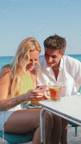 Young couple enjoying a sunny beach day while looking at their smartphones and sharing joyful moments together on the sand by the beautiful coastline with friends nearby on a lively summer afternoon photo