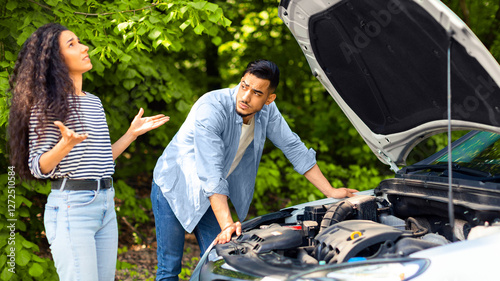 Upset middle-eastern couple having broken car while countryside trip, puzzled arab guy checking engine, emotional brunette lady looking up, sad mixed race family stuck in the middle of road photo